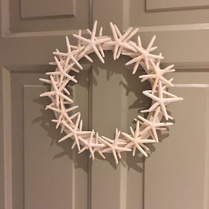 May include: A white starfish wreath with a light brown background. The wreath is made of many starfish that are arranged in a circle.
