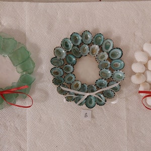 Puede incluir: Tres coronas de Navidad hechas a mano con diferentes materiales. La primera corona está hecha con trozos de vidrio verde, la segunda con conchas verdes y blancas, y la tercera con conchas blancas. Cada corona tiene una cinta roja atada alrededor.
