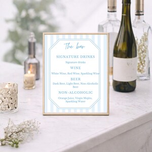 May include: A framed bar menu with a light blue and white striped background. The menu lists signature drinks, wine, beer, and non-alcoholic beverages. The text is in a stylish, elegant font. The frame is gold.