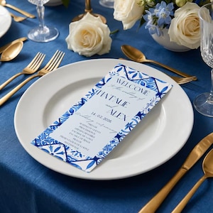 Può includere: Un'impostazione del tavolo nuziale con un piatto bianco, posate dorate e un biglietto di benvenuto blu e bianco. Il biglietto recita "Benvenuti al matrimonio di Natalie e Alex, 14.02.2026." Rose bianche e fiori blu in un vaso.