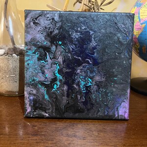 Puede incluir: Arte en lienzo cuadrado con un diseño abstracto. La obra presenta un fondo oscuro con patrones arremolinados de pintura negra, verde azulado y púrpura. El lienzo se exhibe sobre una superficie de madera.