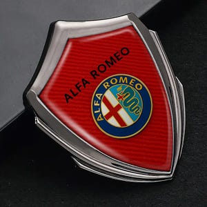May include: A silver-edged emblem featuring the Alfa Romeo logo. The emblem has a red background with the words "ALFA ROMEO" in white. The logo is a circular design with a white cross, a green serpent, and blue and gold accents.
