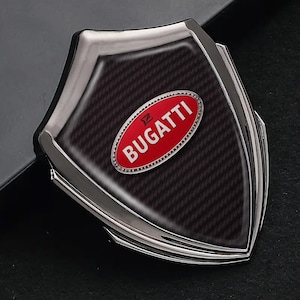Puede incluir: Un llavero de coche en forma de escudo con un patrón de fibra de carbono y un borde plateado. El emblema ovalado rojo en el centro presenta la palabra "BUGATTI" en letras blancas.