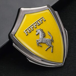 May include: A silver and yellow Ferrari emblem. The emblem features a yellow shield with the word "Ferrari" in silver lettering at the top. A silver horse is in the centre of the shield. The emblem has a silver border.