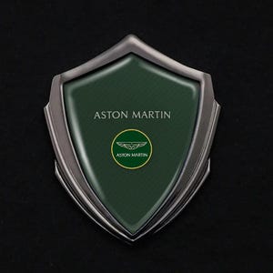 Può includere: Un emblema a forma di scudo con sfondo verde scuro e le parole "ASTON MARTIN" in bianco. L'emblema presenta un logo circolare con un design alato e le parole "ASTON MARTIN" in un cerchio dorato. Lo scudo ha un bordo color argento.