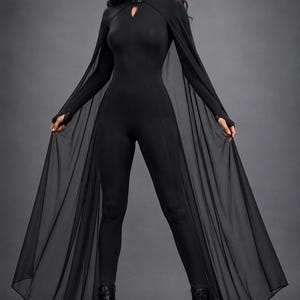 May include: A black long-sleeved jumpsuit featuring a high collar with a small cutout detail at the neckline. The garment includes long, sheer, flowing cape-like panels attached to the shoulders that extend to the floor, paired with black boots.