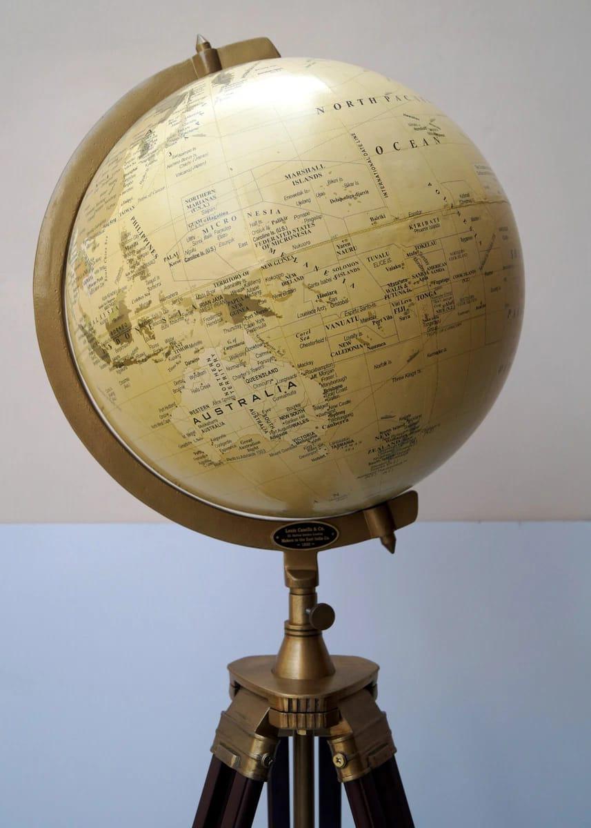 Könnte beinhalten: Ein Globus im Vintage-Stil mit cremefarbener Oberfläche, der geografische Details und Ländernamen zeigt. Der Globus ist auf einem messingfarbenen Ständer mit einem Dreibein-Sockel montiert. Der Ständer hat dunkelbraune Holzbeine. Ein dekoratives und lehrreiches Objekt.
