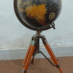 Op de afbeelding: Een wereldbol in vintage-stijl met een zwart-gouden kleurenschema, gemonteerd op een houten statief. De wereldbol toont gedetailleerde geografische markeringen en wordt ondersteund door een driepootstandaard met metalen accenten. Het ontwerp heeft een klassieke, antieke uitstraling.