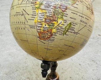 Op de afbeelding: Een vintage wereldbol met een crèmekleurig oppervlak, waarop continenten en landen in verschillende kleuren worden weergegeven. De wereldbol is gemonteerd op een donkerbronzen standaard met een driepoot, ideaal voor decoratie.