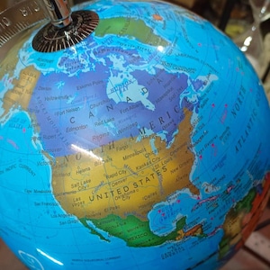 Op de afbeelding: Een gedetailleerde wereldbol met een blauwe oceaan en kleurrijke continenten. De wereldbol toont de Verenigde Staten, Canada en Mexico, met stadsnamen en geografische details. Een zilveren metalen meridiaan en voetstuk maken het ontwerp compleet.