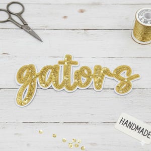 Könnte beinhalten: Ein goldfarbener Pailletten-Aufnäher "Gators" mit weißem Rand, auf einer weißen Holzoberfläche. Nähutensilien, darunter eine Schere, goldfarbenes Garn und ein "HANDMADE"-Etikett sind zu sehen. Streupailletten ergänzen das Bild.