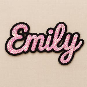 Op de afbeelding: Een roze pailletten patch met de naam "Emily" in sierlijke letters, omlijnd met zwart. Geschikt voor kleding of accessoires, tegen een neutrale achtergrond.