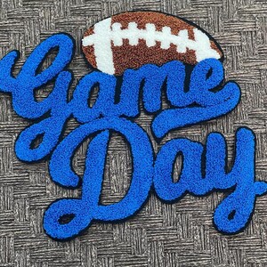 Puede incluir: Un diseño azul texturizado de "Game Day" con un gráfico de fútbol americano. El texto está en cursiva y el balón de fútbol es marrón con rayas blancas. El diseño es probablemente para los fanáticos del deporte.