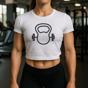 May include: White crop top with a black outline of a kettlebell and barbell design. The short-sleeved top has a crew neck. The image is taken in a gym setting.