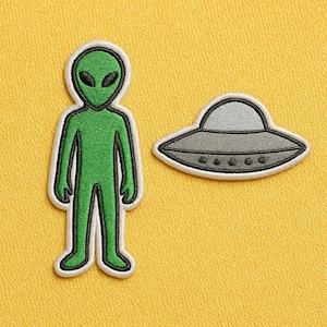 Op de afbeelding: Twee geborduurde patches: een groene alien met zwarte ogen en een grijze UFO. De alien is zwart-wit omlijnd en staat rechtop. De UFO is grijs met zwarte details en een witte rand. Beide patches staan op een gele achtergrond.