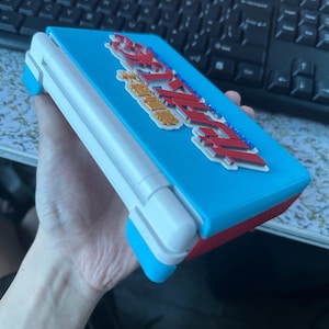 May include: A light blue handheld gaming console with a white hinge and a red base. The top cover features a raised, multi-coloured graphic logo with Japanese text in red, white, and blue, displaying the characters for 'Rockman' or Mega Man.