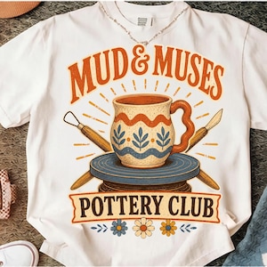 May include: A white t-shirt featuring a graphic print of a ceramic mug on a pottery wheel, flanked by two sculpting tools. The text reads MUD & MUSES POTTERY CLUB in orange and black, accented by small floral illustrations at the bottom.