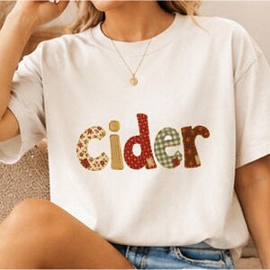 Puede incluir: Una camiseta de manga corta color crema con la palabra cider impresa en el pecho. Cada letra tiene un patrón distinto: la c es floral, la i es dorada, la d tiene lunares rojos, la e es de cuadros verdes y la r es floral marrón.
