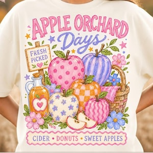 Può includere: Una t-shirt color crema con una grafica che riporta il testo APPLE ORCHARD Days e CIDER DONUTS SWEET APPLES. Il design include un cartello in legno con l'etichetta FRESH PICKED, una caraffa di sidro, un cesto di vimini e cinque mele decorate.