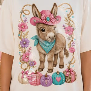 May include: A cream-colored t-shirt featuring a graphic of a brown donkey wearing a pink cowboy hat and a turquoise bandana. The donkey is surrounded by pink and purple flowers, rope accents, and four decorative pumpkins at the bottom.