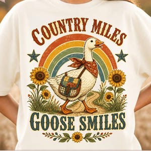 Puede incluir: Una camiseta color crema con el gráfico de un ganso blanco que lleva un pañuelo rojo estampado y un bolso de retazos. El ganso está sobre un camino de tierra frente a un arcoíris, girasoles y estrellas, con el texto Country Miles Goose Smiles.
