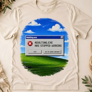 Könnte beinhalten: Ein beiges Kurzarm-T-Shirt mit einer kreisförmigen Grafik aus blauem Himmel und grüner Hügellandschaft. Zentriert ist ein graues Computer-Fehlerfenster mit dem Text Adulting.exe, ADULTING.EXE HAS STOPPED WORKING sowie den Schaltflächen CRY und GO TO WORK ANYWAY.