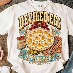 Könnte beinhalten: Ein weißes T-Shirt mit einem Grafikdruck, der eine Platte mit gefüllten Eiern, eine rote Schleife, eine mit PAPRIKA beschriftete Dose, eine Gabel und eine rot karierte Serviette zeigt. Der Text DEVILED EGG DEPARTMENT ist in gold-blauer Schrift.
