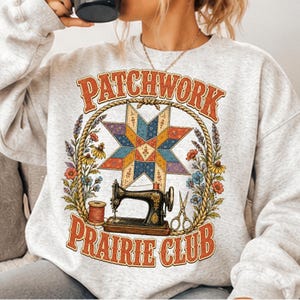 May include: A light gray crewneck sweatshirt featuring a graphic print with the text "PATCHWORK PRAIRIE CLUB" in orange. The design includes a colorful star-shaped quilt block, a black sewing machine, a spool of thread, scissors, and flowers.