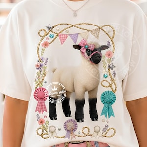 May include: A cream-coloured t-shirt featuring a graphic of a lamb with black legs and face, wearing a pink plaid bow. The lamb is framed by a rope border with flowers, bunting, and award ribbons. The text ClipCountryClub is visible.