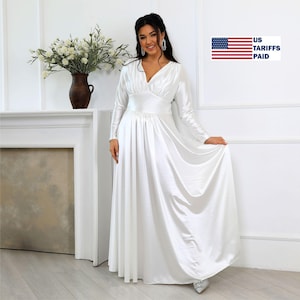 May include: A long, white satin dress with long sleeves and a V-neck. The dress has a fitted bodice and a flowing skirt. The image also includes a vase of white flowers and the text "US Tariffs Paid".