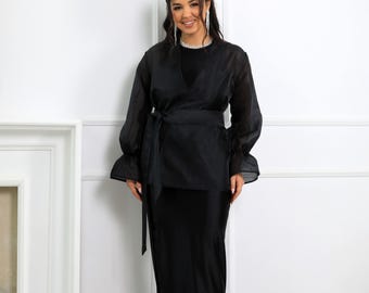May include: A black outfit comprising a sheer, long-sleeved wrap top with a tie belt and a long, form-fitting skirt. The top has ruffled cuffs. The outfit is worn by a person.