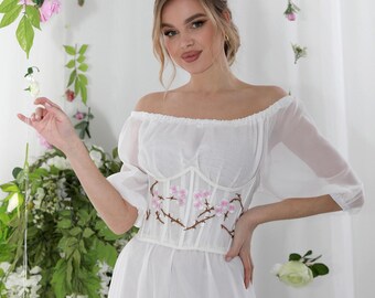 May include: A white off-the-shoulder dress with a floral embroidered bodice. The bodice features a delicate design of pink flowers and brown stems. The dress has sheer sleeves and a flowing skirt, creating an elegant and romantic look.