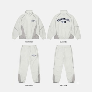 Peut inclure: Un survêtement blanc et gris clair composé d'une veste zippée et d'un pantalon assorti. La veste présente un col montant et le texte bleu foncé CUSTOM LOGO TEXT sur la poitrine et le dos. Le pantalon porte le texte bleu foncé sur la cuisse.