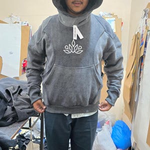 May include: A gray hoodie with a large hood and a front pocket. The hoodie has a white floral design on the chest and a tag attached to the collar. The person is wearing black pants and a white shirt underneath.