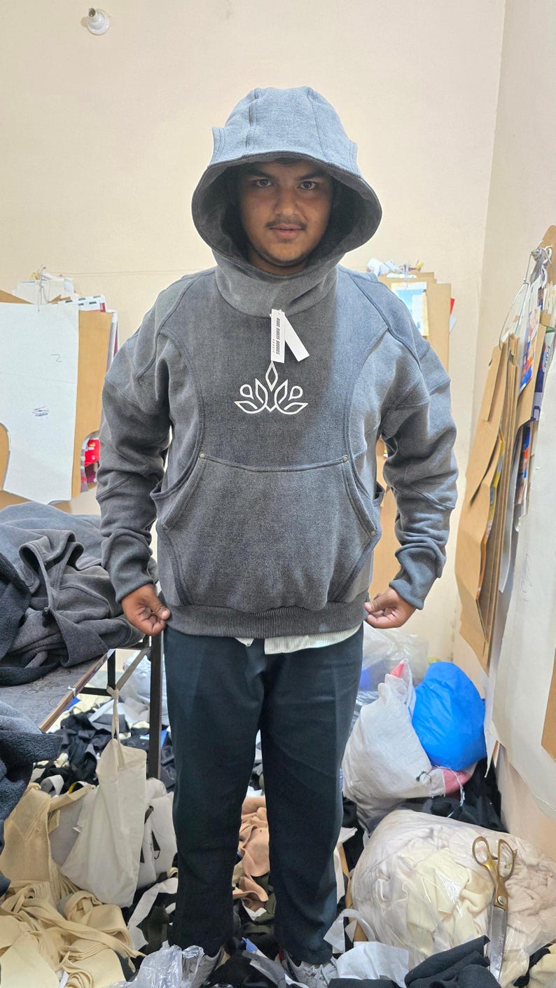 May include: A gray hoodie with a large hood and a front pocket. The hoodie has a white floral design on the chest and a tag attached to the collar. The person is wearing black pants and a white shirt underneath.