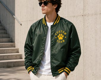 May include: A dark green satin bomber jacket with yellow striped ribbed trim on the collar, cuffs, and hem. The jacket features a yellow graphic on the left chest displaying a paw print icon surrounded by the text CUSTOM TEAM and two stars.