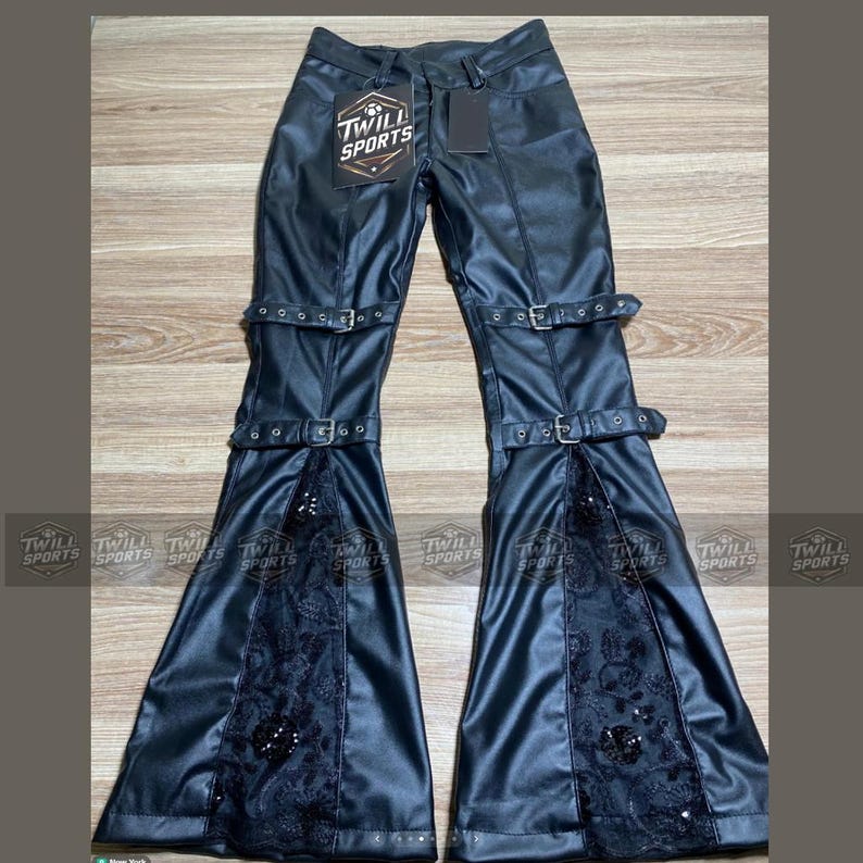 Puede incluir: Pantalones negros de cuero sint&eacute;tico con perneras acampanadas, con hebillas decorativas y paneles de encaje con lentejuelas. Los pantalones tienen el texto "TWILL SPORTS" en una etiqueta.