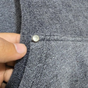 May include: Close-up of a gray hooded sweatshirt with a silver button detail. The fabric has a textured appearance. A white embroidered design is visible on the upper right side of the sweatshirt.