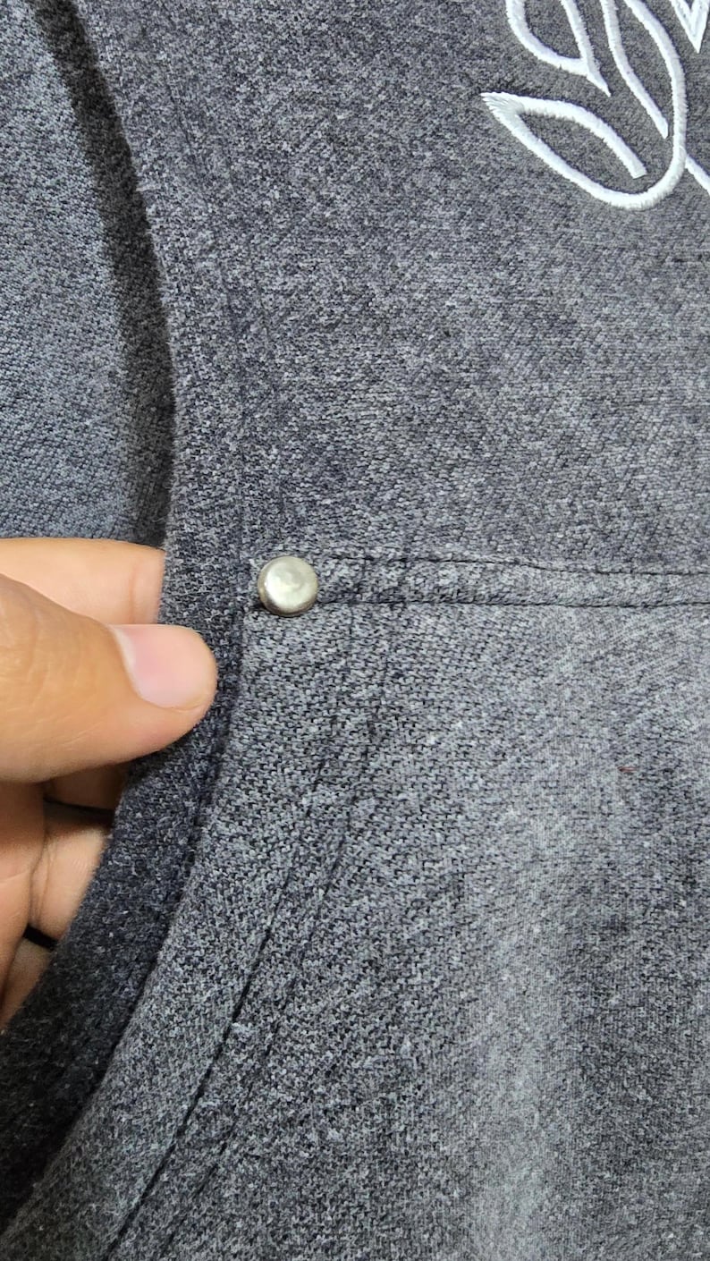 May include: Close-up of a gray hooded sweatshirt with a silver button detail. The fabric has a textured appearance. A white embroidered design is visible on the upper right side of the sweatshirt.