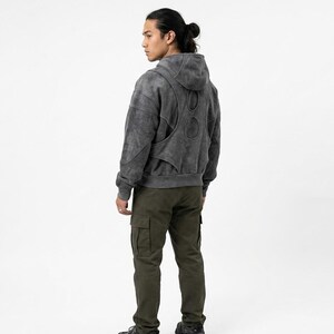 May include: A person wearing a gray hooded sweatshirt with a unique cut-out design on the back and olive green cargo pants. The sweatshirt has a faded, textured appearance. The pants have multiple pockets.