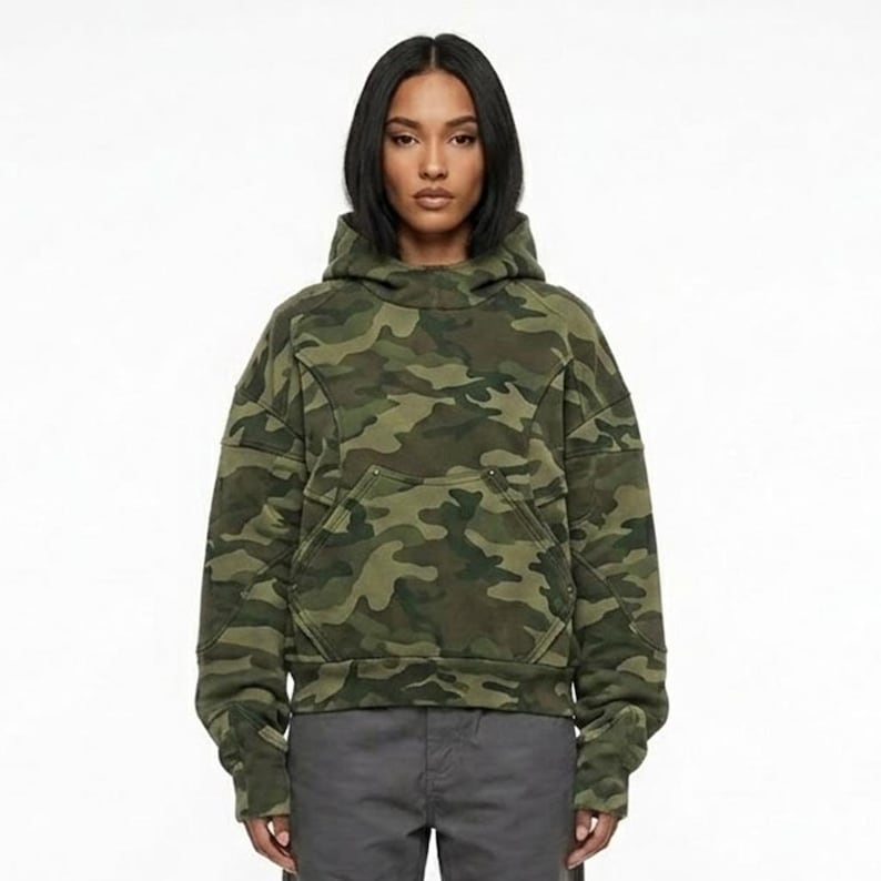 May include: A green camouflage hoodie with a front pocket and a high collar. The hoodie has a relaxed fit and is made of a soft material. The camouflage pattern features shades of green, brown, and black.