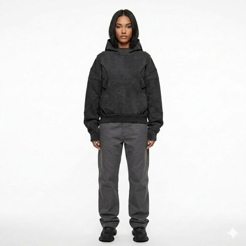 May include: A person wearing a dark gray hoodie with a front pocket and matching gray pants. The hoodie has a unique design with seams and a hood. The pants are straight-legged and the person is wearing black shoes.