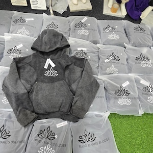May include: A gray hooded sweatshirt with a white logo is displayed on top of several gray plastic bags. The bags are printed with a logo and text. The sweatshirt has a kangaroo pocket.