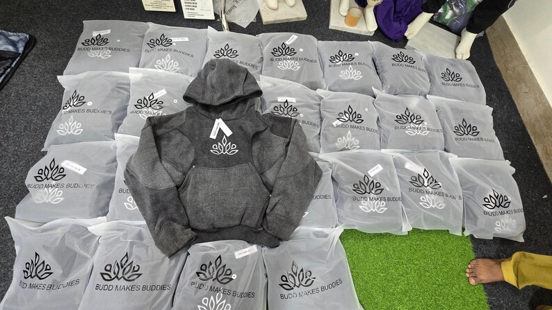 May include: A gray hooded sweatshirt with a white logo is displayed on top of several gray plastic bags. The bags are printed with a logo and text. The sweatshirt has a kangaroo pocket.