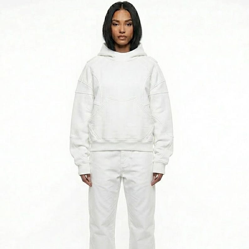 May include: A white hoodie with a matching pair of white pants. The hoodie has a front pocket and stitched detailing. The model is wearing a white outfit against a white background.