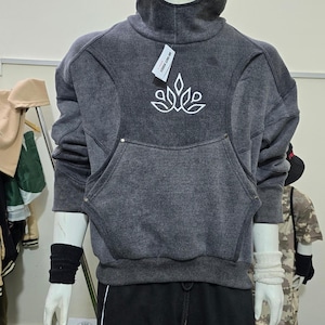 May include: A dark gray hoodie with a large front pocket and a white floral design. The hoodie has a high collar and a drawstring hood. The mannequin is also wearing black sweatpants with a white stripe.