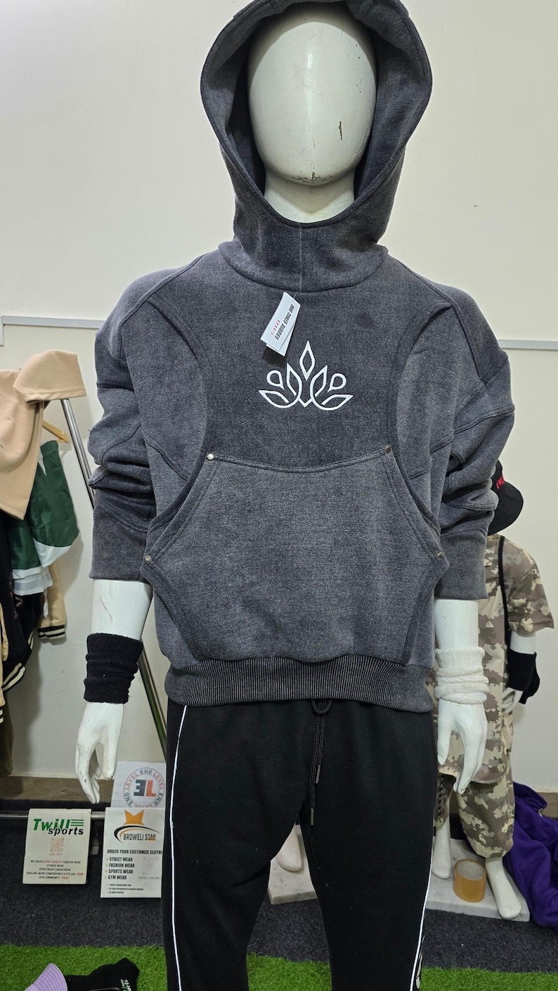 May include: A dark gray hoodie with a large front pocket and a white floral design. The hoodie has a high collar and a drawstring hood. The mannequin is also wearing black sweatpants with a white stripe.