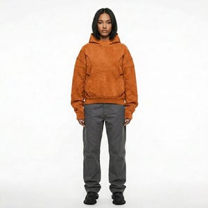 May include: A person wearing a rust-colored hoodie with a kangaroo pocket and gray pants. The hoodie has a textured appearance and a drawstring hood. The pants are a solid gray color and have a straight leg.
