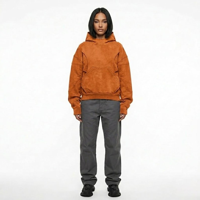 May include: A person wearing a rust-colored hoodie with a kangaroo pocket and gray pants. The hoodie has a textured appearance and a drawstring hood. The pants are a solid gray color and have a straight leg.