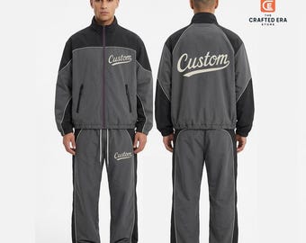 Può includere: Una tuta grigio antracite e nera composta da una giacca a collo alto e pantaloni abbinati. La giacca presenta una cerniera frontale, maniche nere e la scritta Custom in corsivo bianco. I pantaloni hanno una vita con coulisse regolabile.